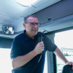 Andrew Donaldson speaks at the front of a bus with a microphone in his hand