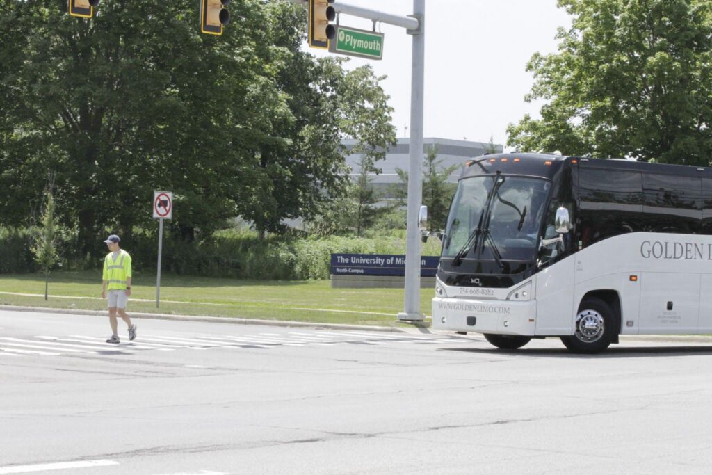 Tinghan Wang, in a yellow vest, walks across a street with a large passenger bus in the background